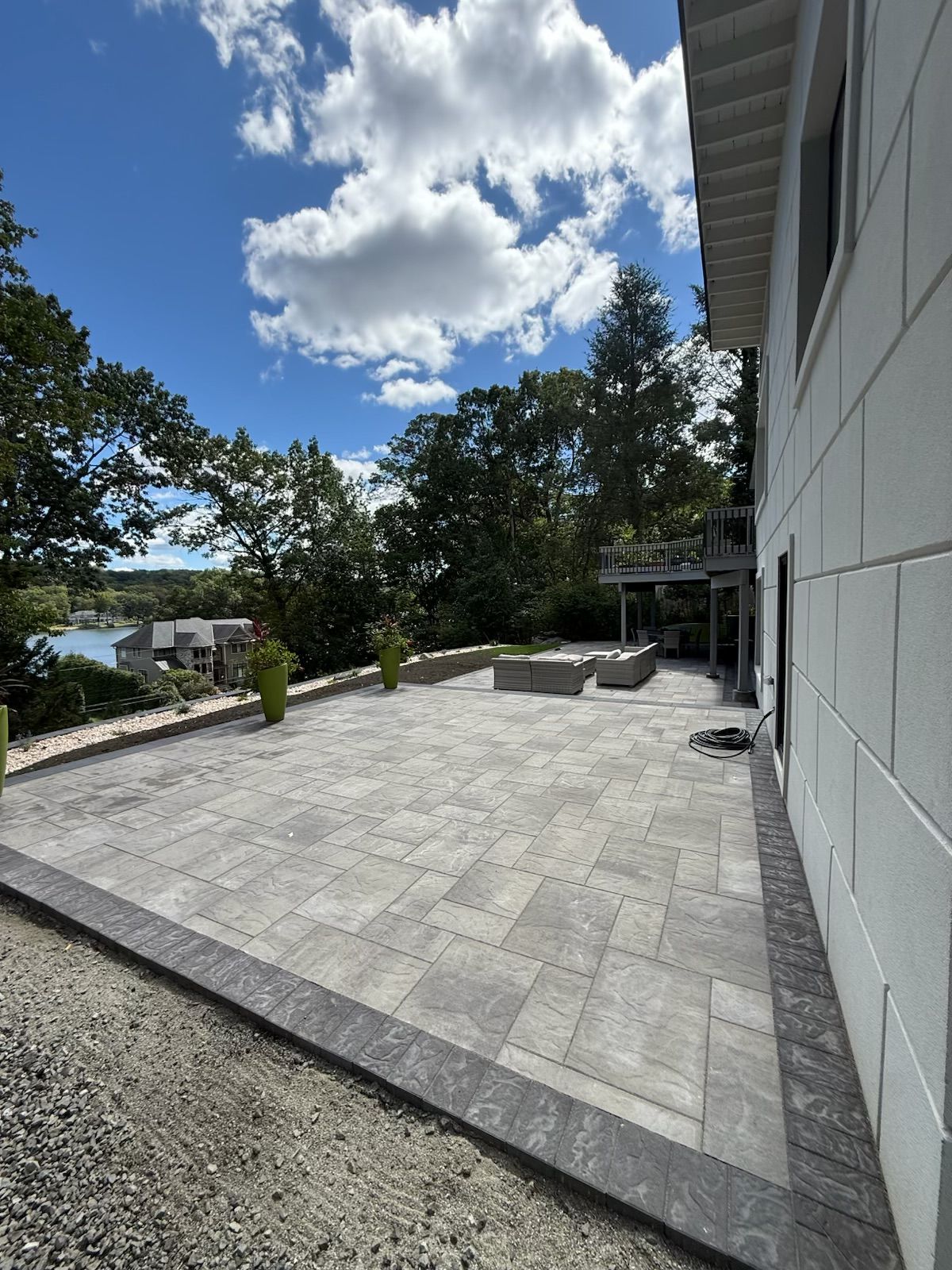 Patio with gray pavers, adjacent to a white building, overlooking a body of water. Sunny, blue sky with clouds.