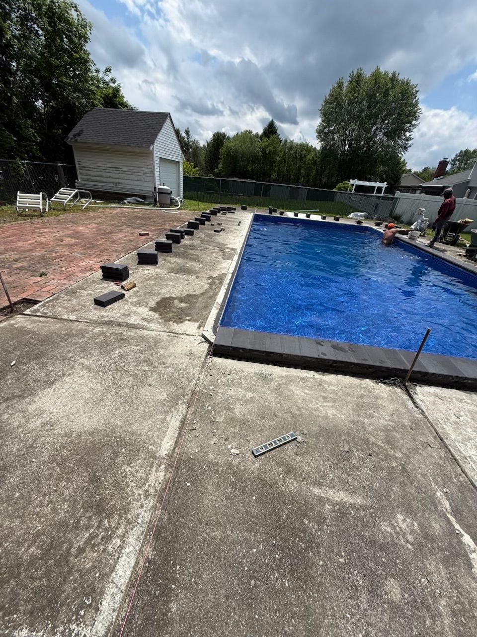 Concrete pool deck with dark border being installed. Cloudy sky, house in background.