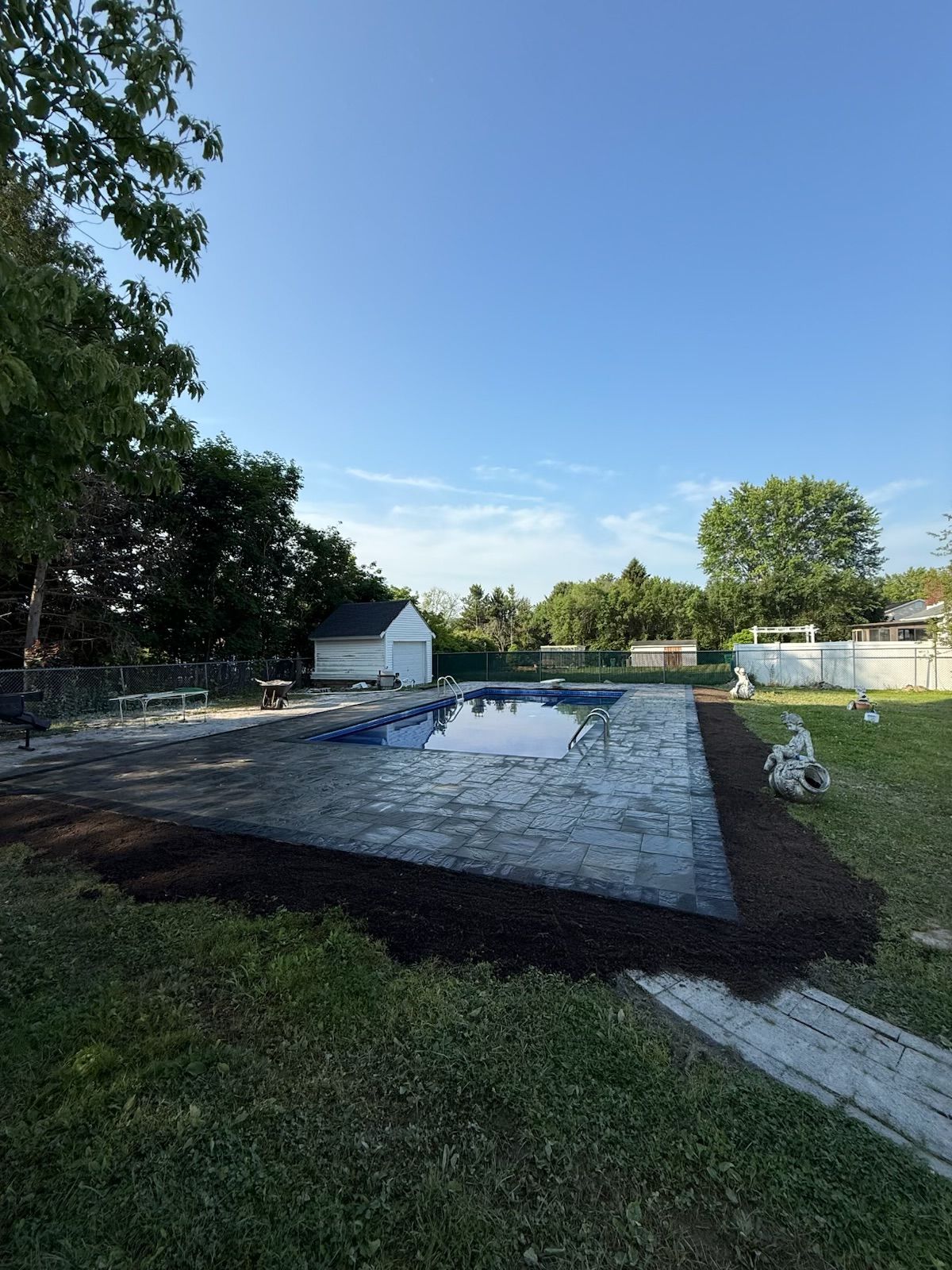 Backyard pool with stone patio, grass, small shed, and landscaping under a blue sky.