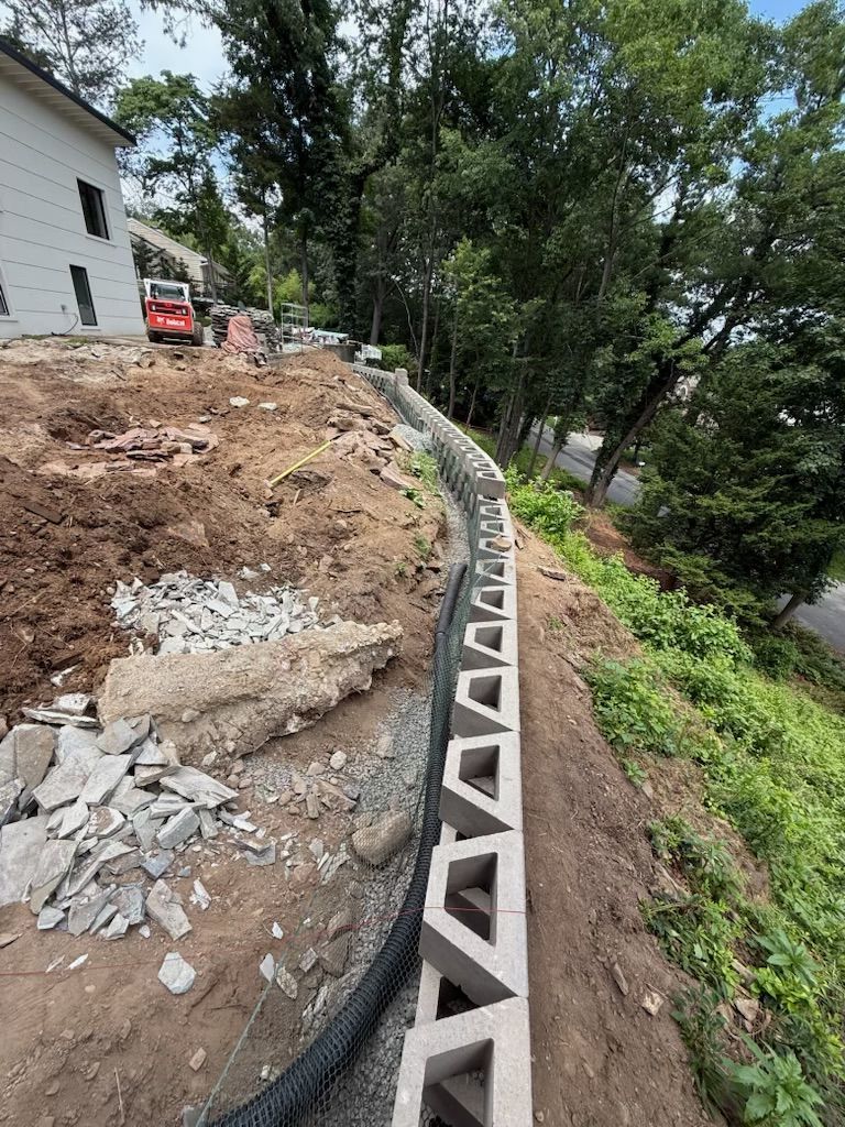 A concrete block retaining wall under construction on a hillside, with a house and trees in the background.