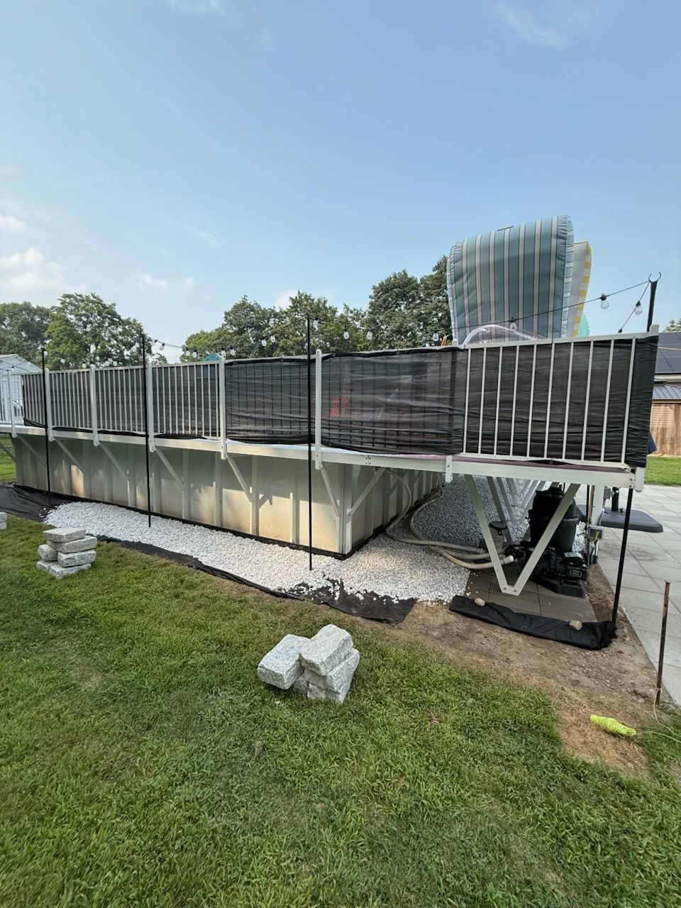 Above-ground pool with black mesh fencing, white gravel border, and green lawn under a blue sky.