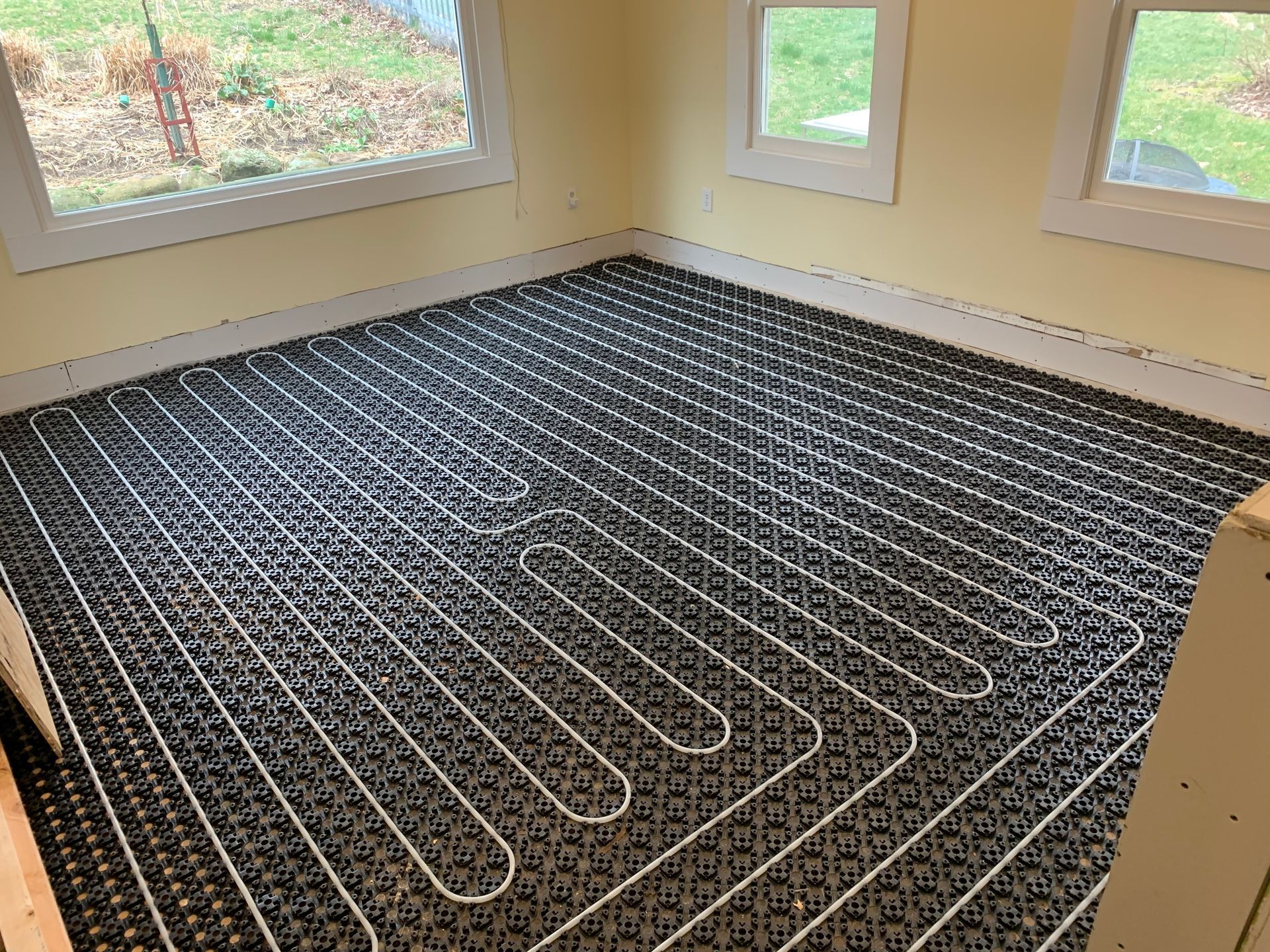 Installed radiant floor heating system in a room with windows; black matting with white tubing laid out.