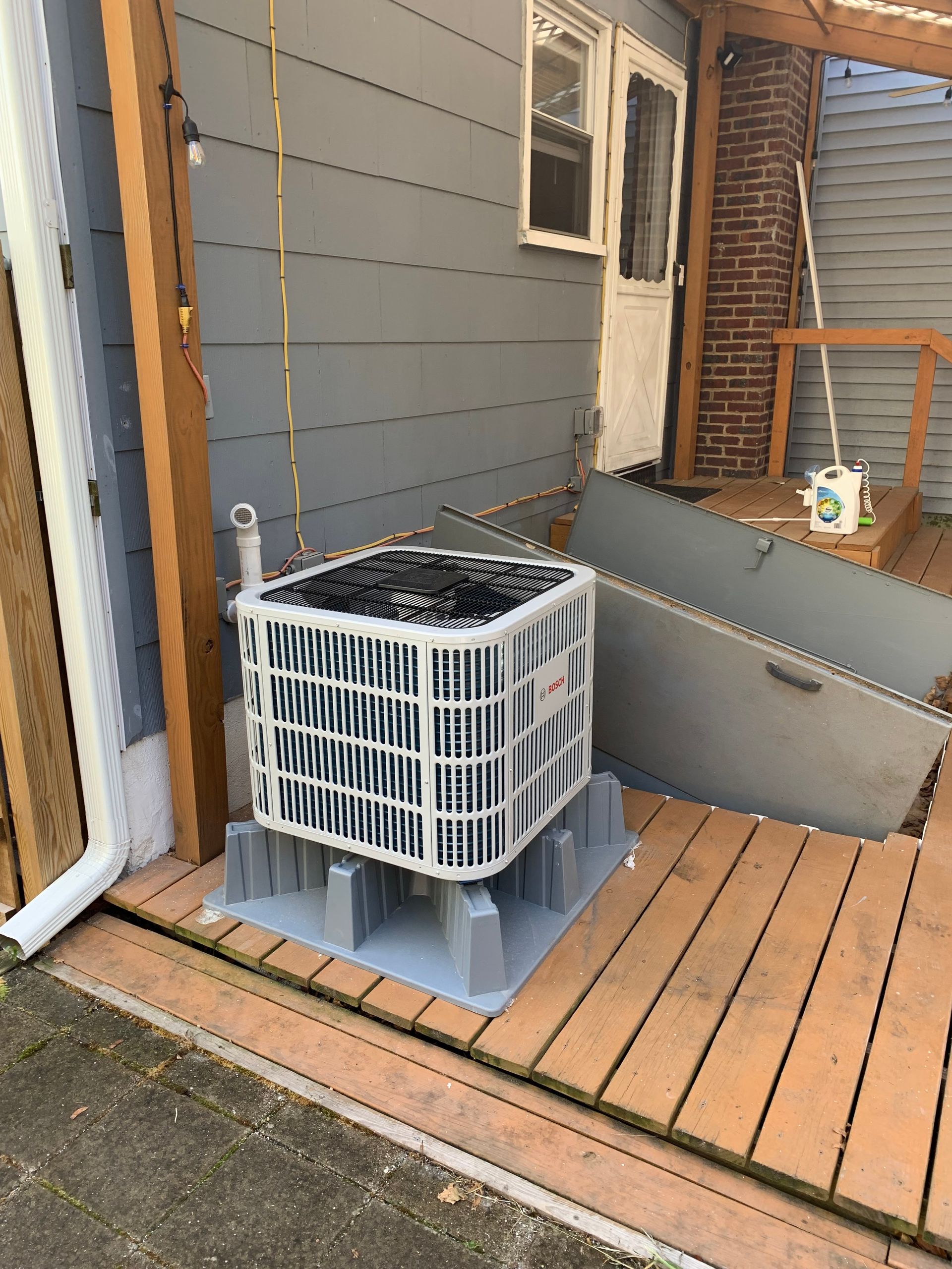 Air conditioning unit on a gray plastic base on a wooden deck next to a house.