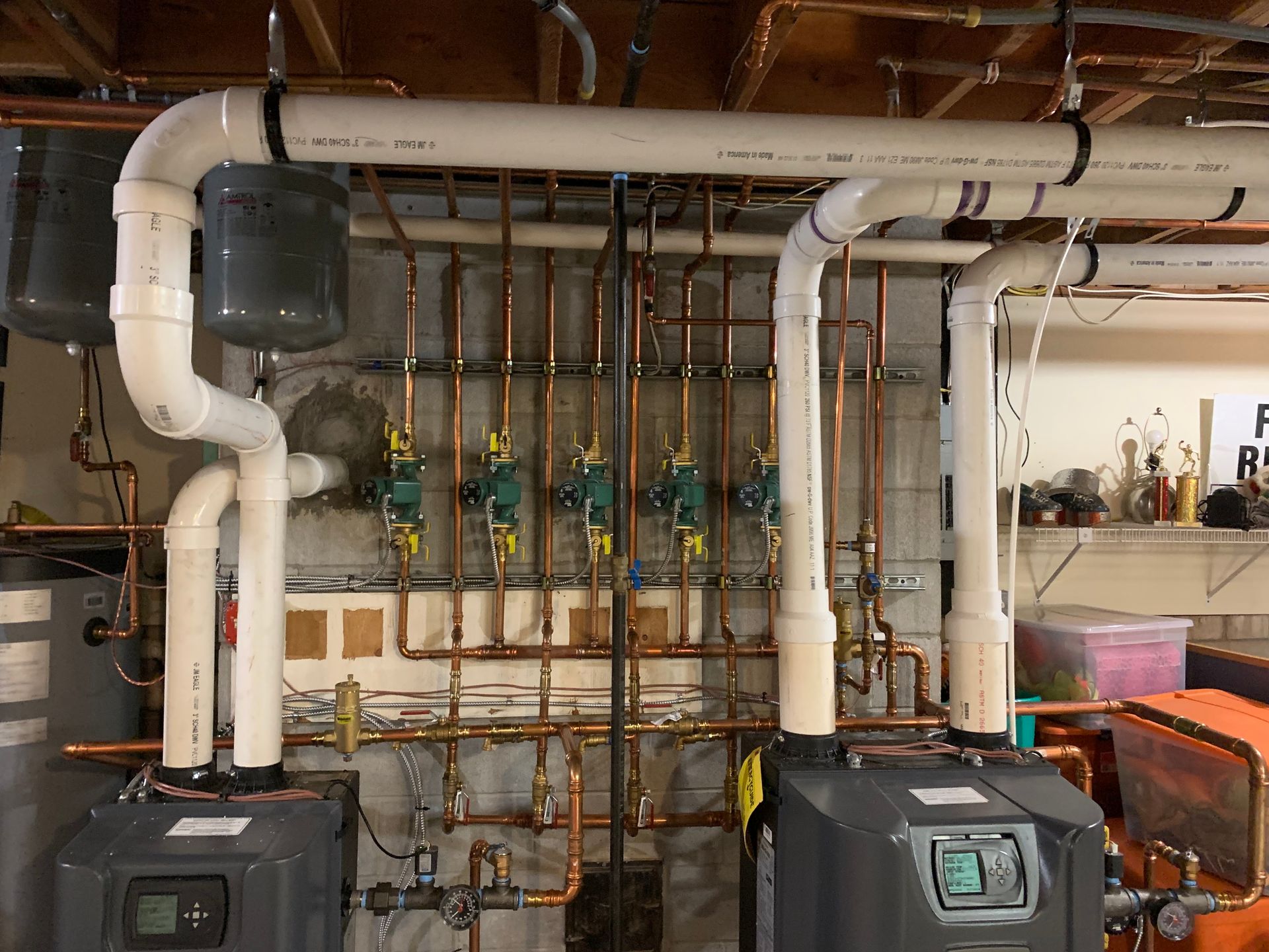 Copper pipes and gray heating units in a utility room; white PVC pipes run overhead.