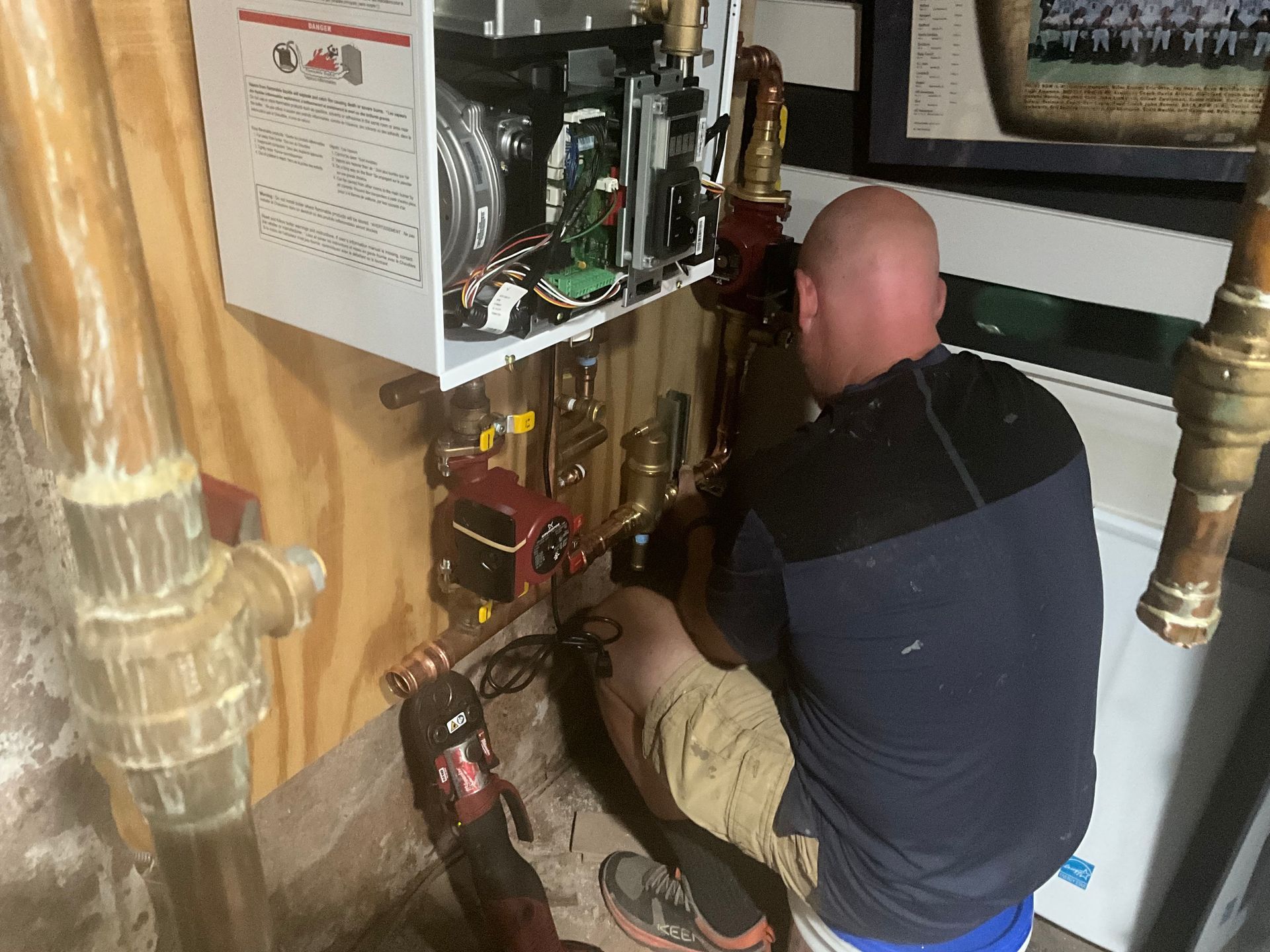 A person working on plumbing in a utility room. They're crouched near copper pipes and a heating unit.