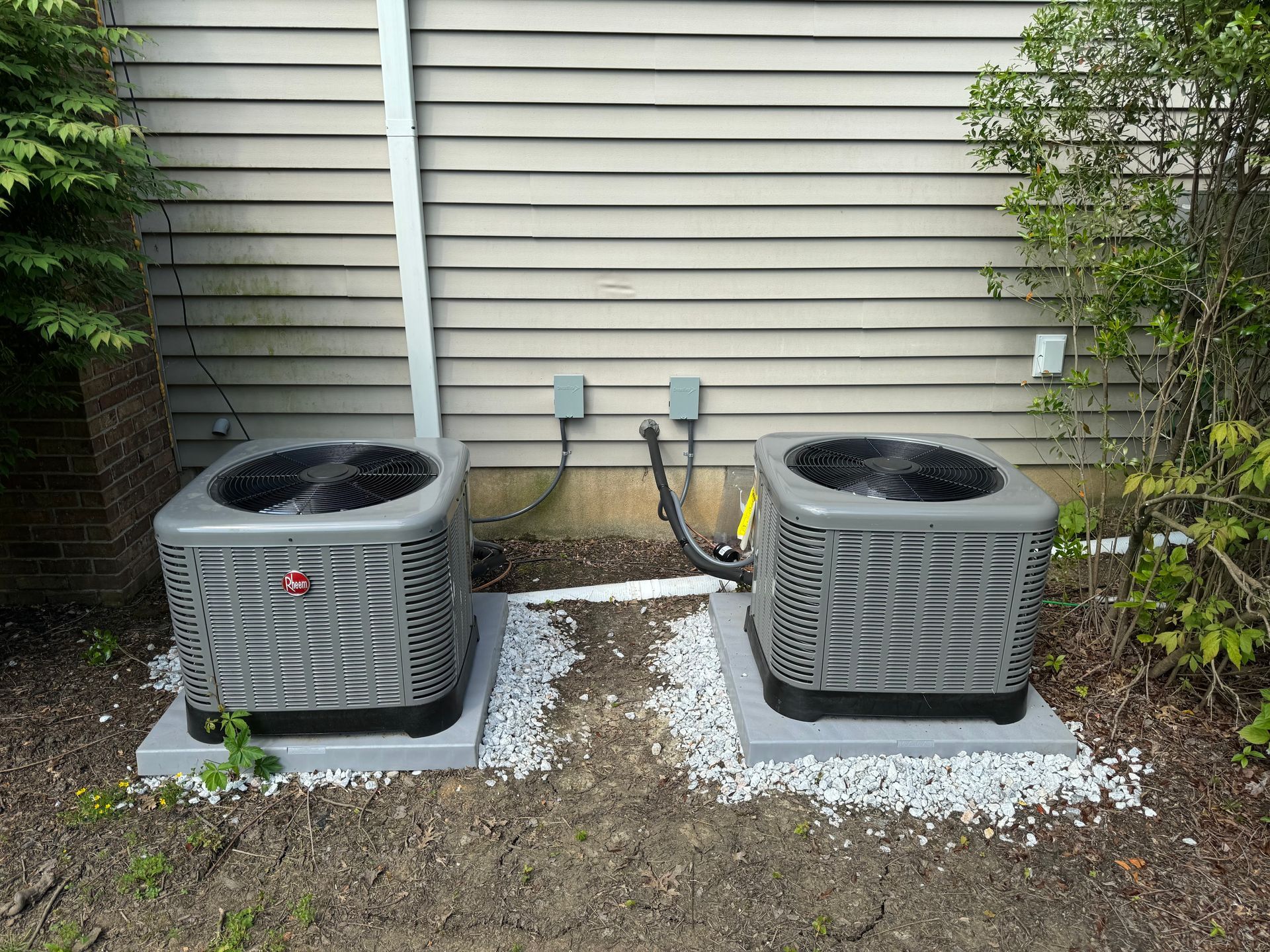 Two grey air conditioning units on concrete pads beside a light brown wall.