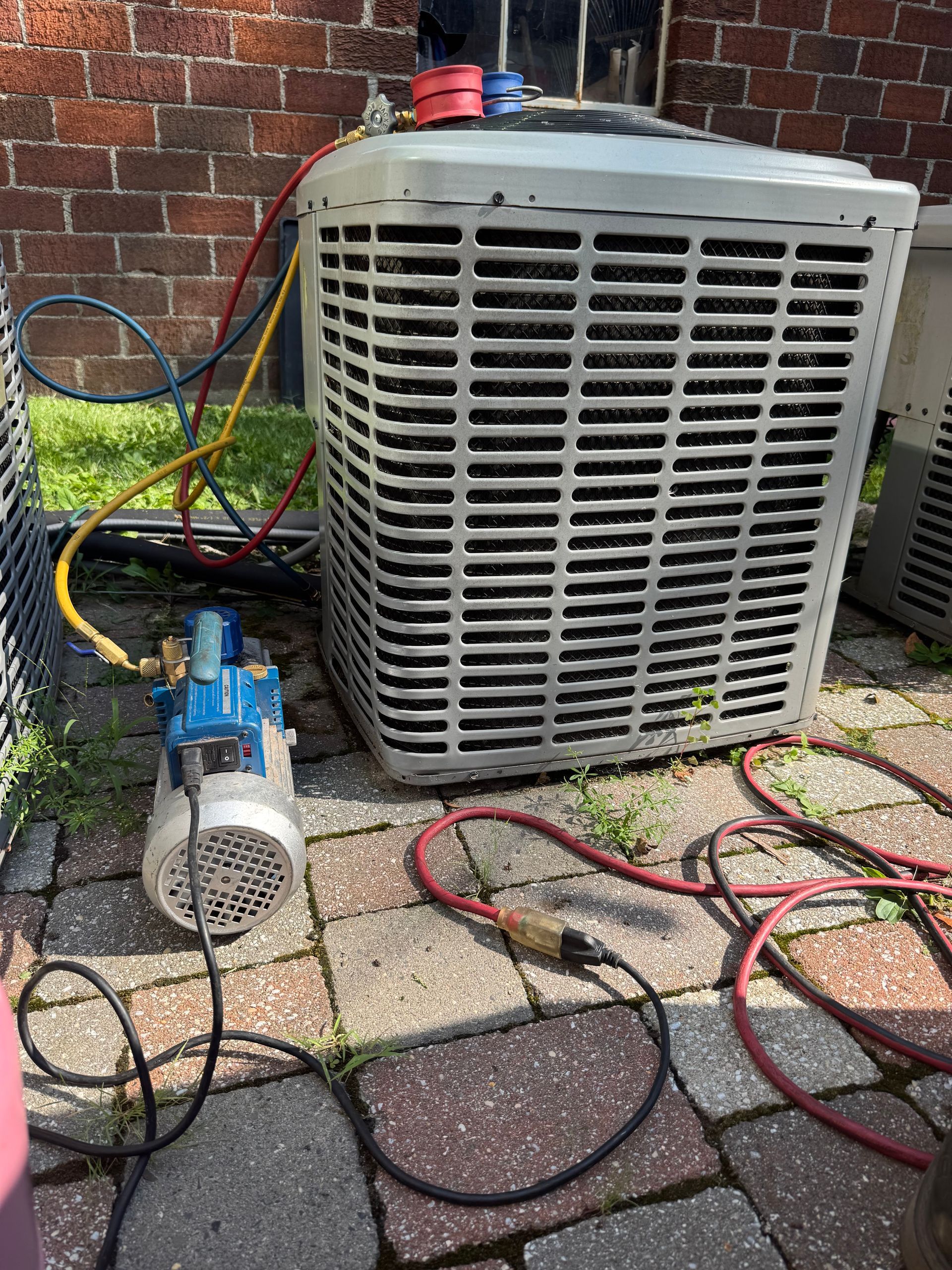 HVAC unit being serviced outdoors, hoses connected to a vacuum pump, against a brick wall.