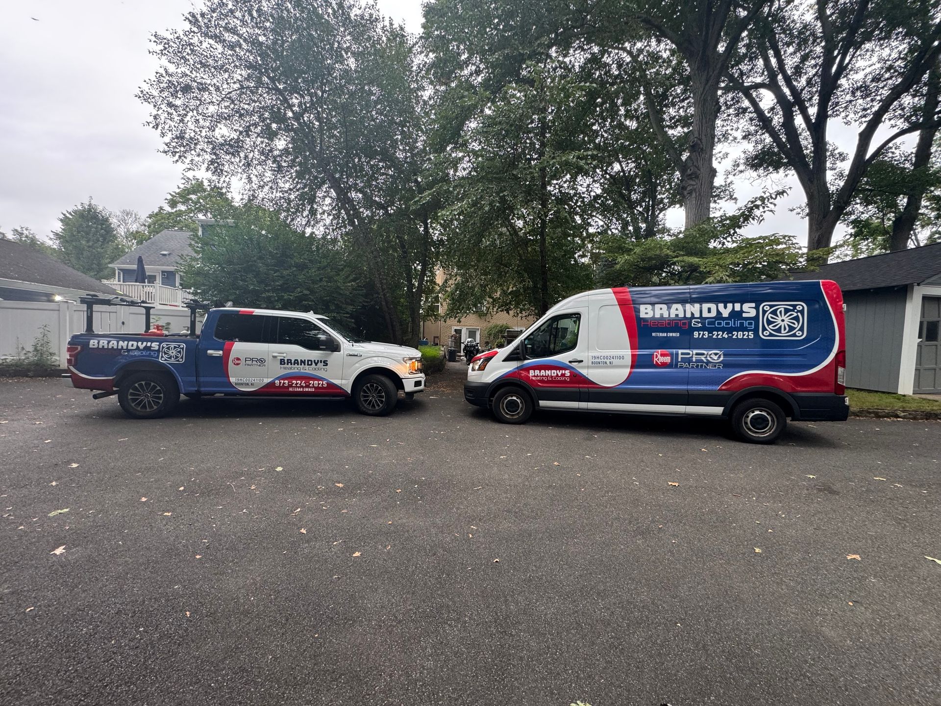 Two vehicles, a truck and a van, with company branding parked on a paved surface. Trees and a house in the background.