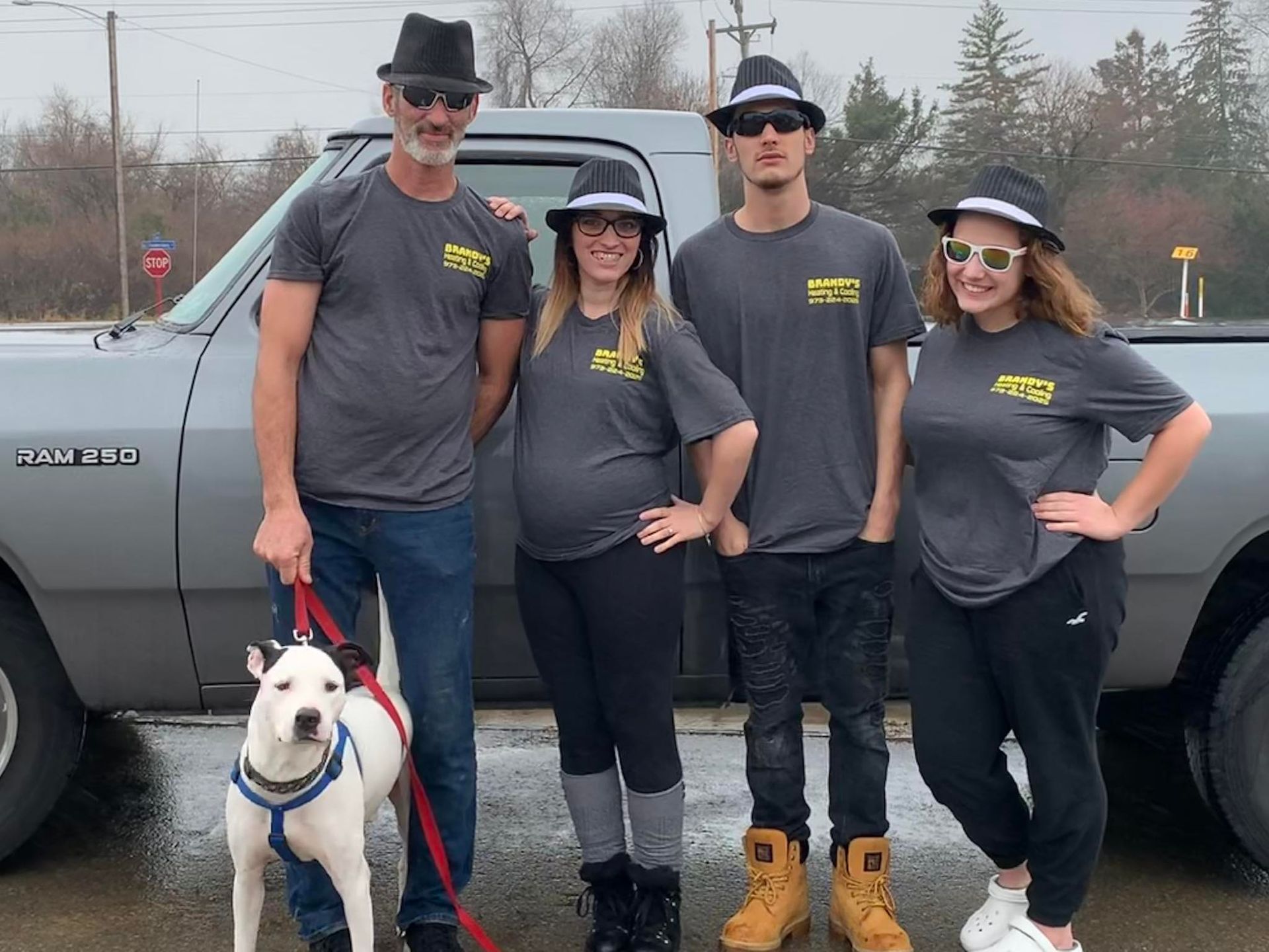 Four people and a dog wearing matching hats and shirts posing next to a truck.