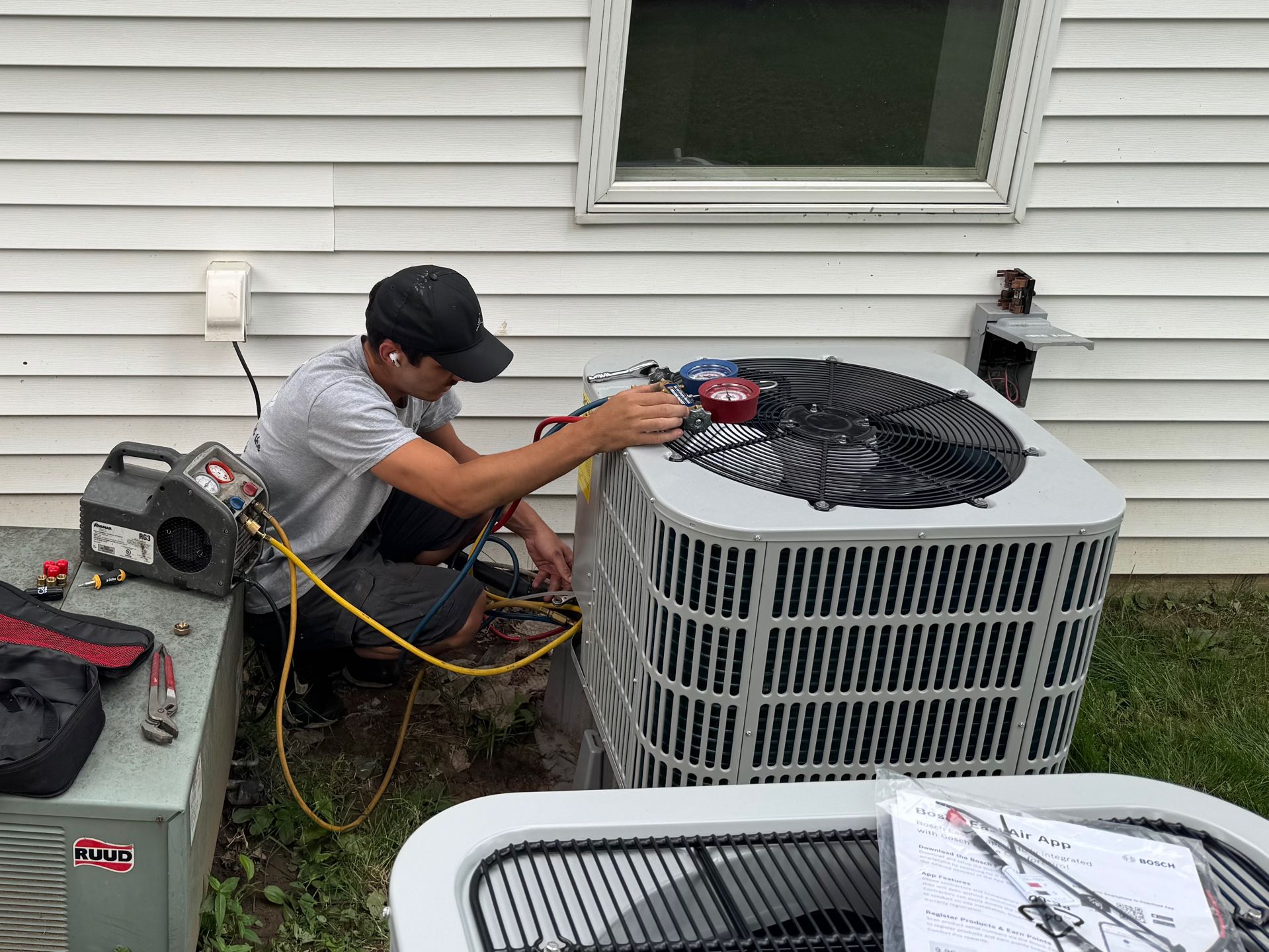 HVAC technician working on an air conditioning unit outside a house. He is kneeling, using tools, and has gauges attached.