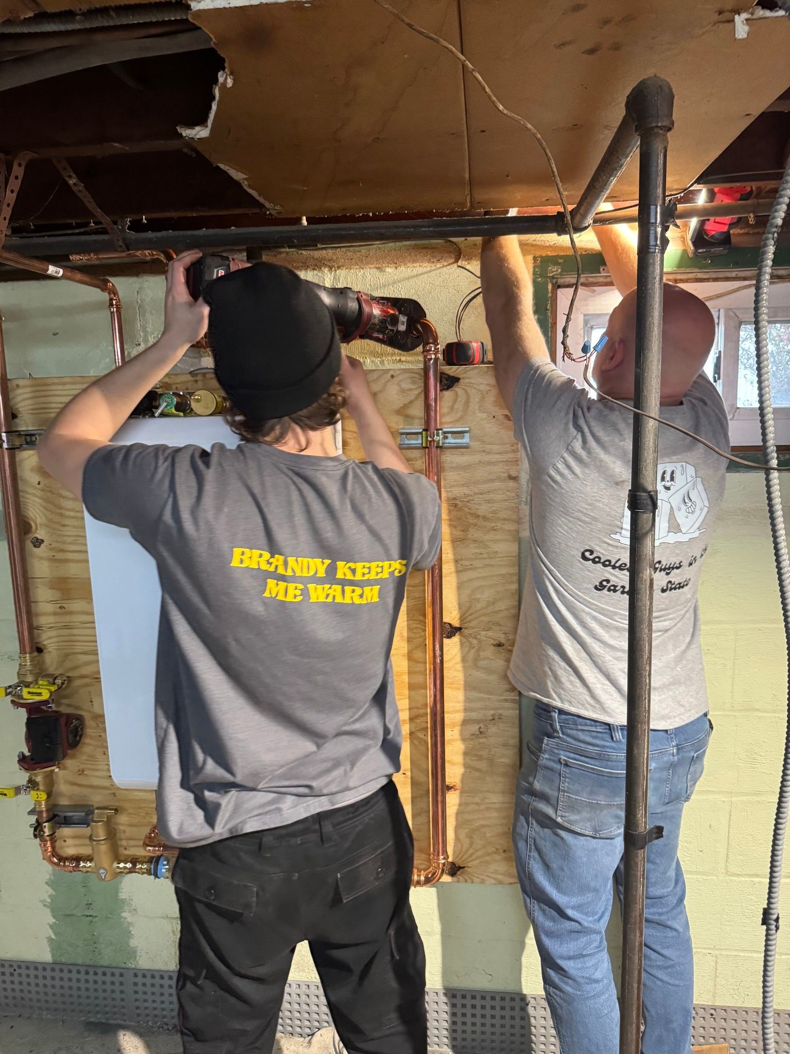 Two people working on pipes in a basement. One uses a power tool, the other works with wires.