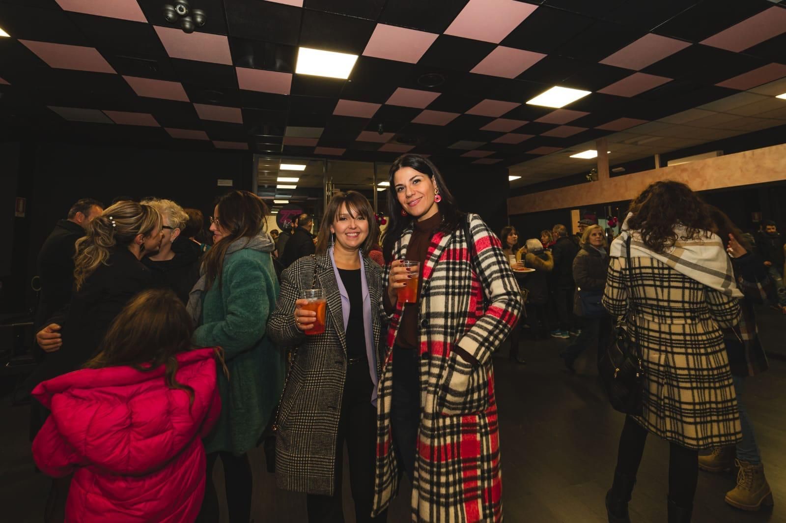 Due donne sono in piedi una accanto all'altra, tenendo in mano dei drink, in una stanza affollata.