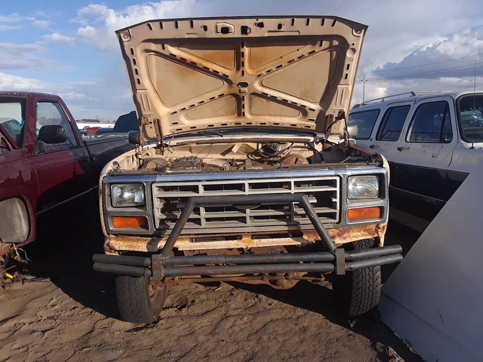 Tan, rusted pickup truck with hood up in junkyard, surrounded by other vehicles.