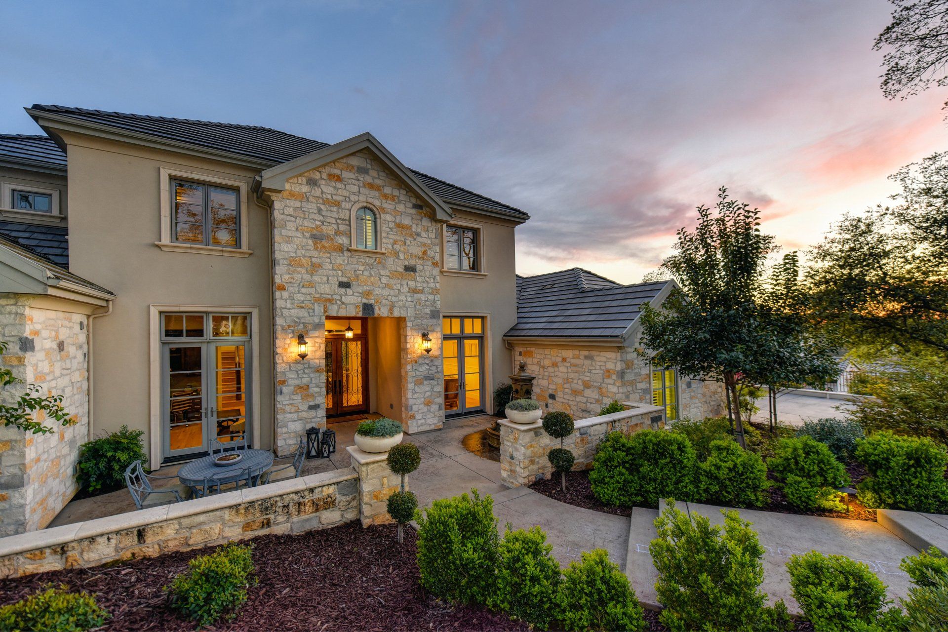 Two-story stone house with tiled roof, lit entryway, and manicured landscaping under a sunset sky.