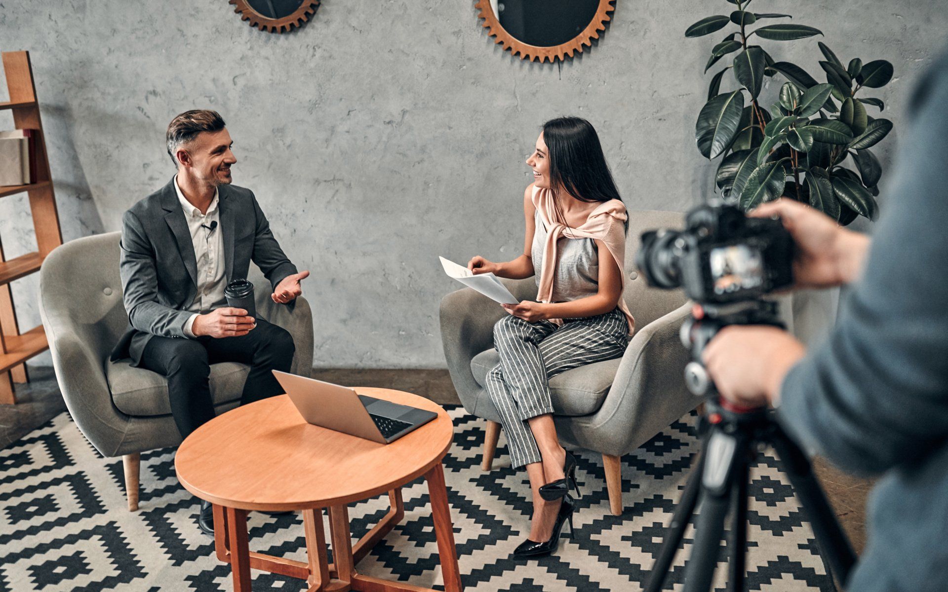 Man being interviewed by a woman in a studio. They sit in chairs with a laptop on a table. A person holds a camera.