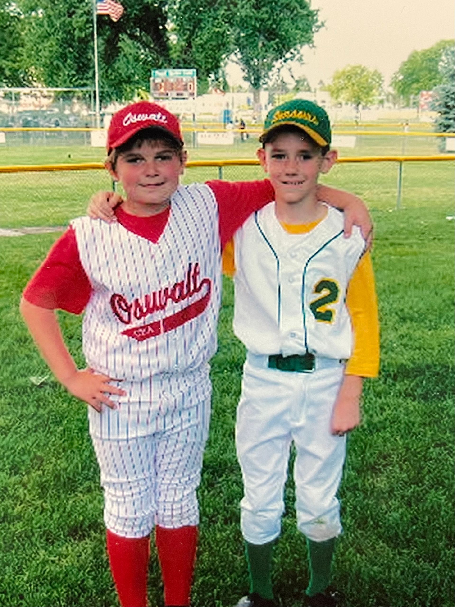 Two little league players in uniform, one with a red cap/uniform, the other with a green cap/uniform, posing on a baseball field.