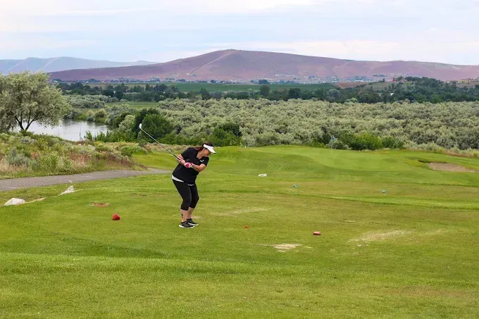 Person in black outfit about to swing a golf club on a green course with a lake and mountain in the background.