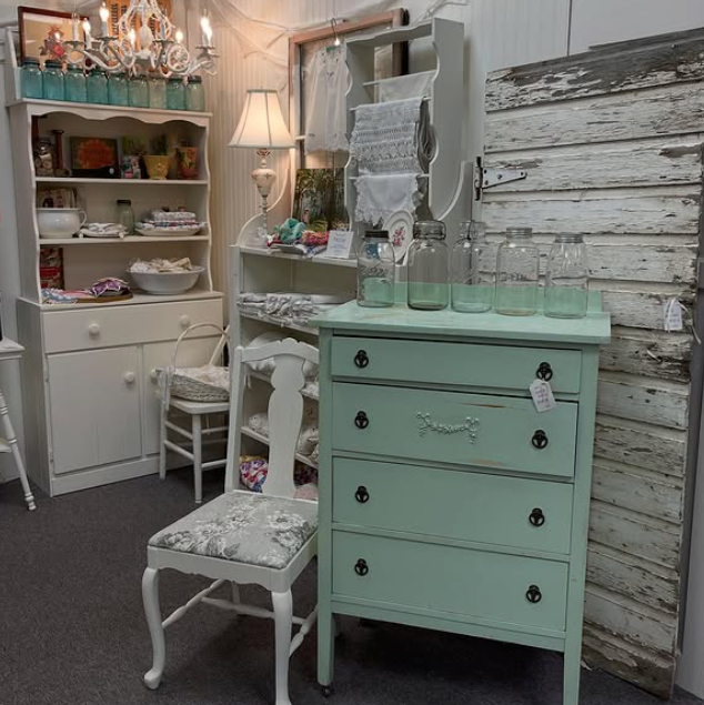 Antique furniture display in a shop, featuring a painted green dresser, white cabinets, and floral-print chair.