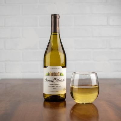 Bottle of Chateau Ste. Michelle Chardonnay next to a glass of wine, on a wooden table. White brick background.