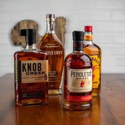 Four whiskey bottles on a wooden table: Knob Creek, Angel's Envy, Pendleton, and Fireball.