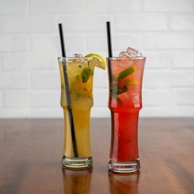 Two tall cocktails with straws on a wooden table, one yellow, one red, in front of a white brick wall.