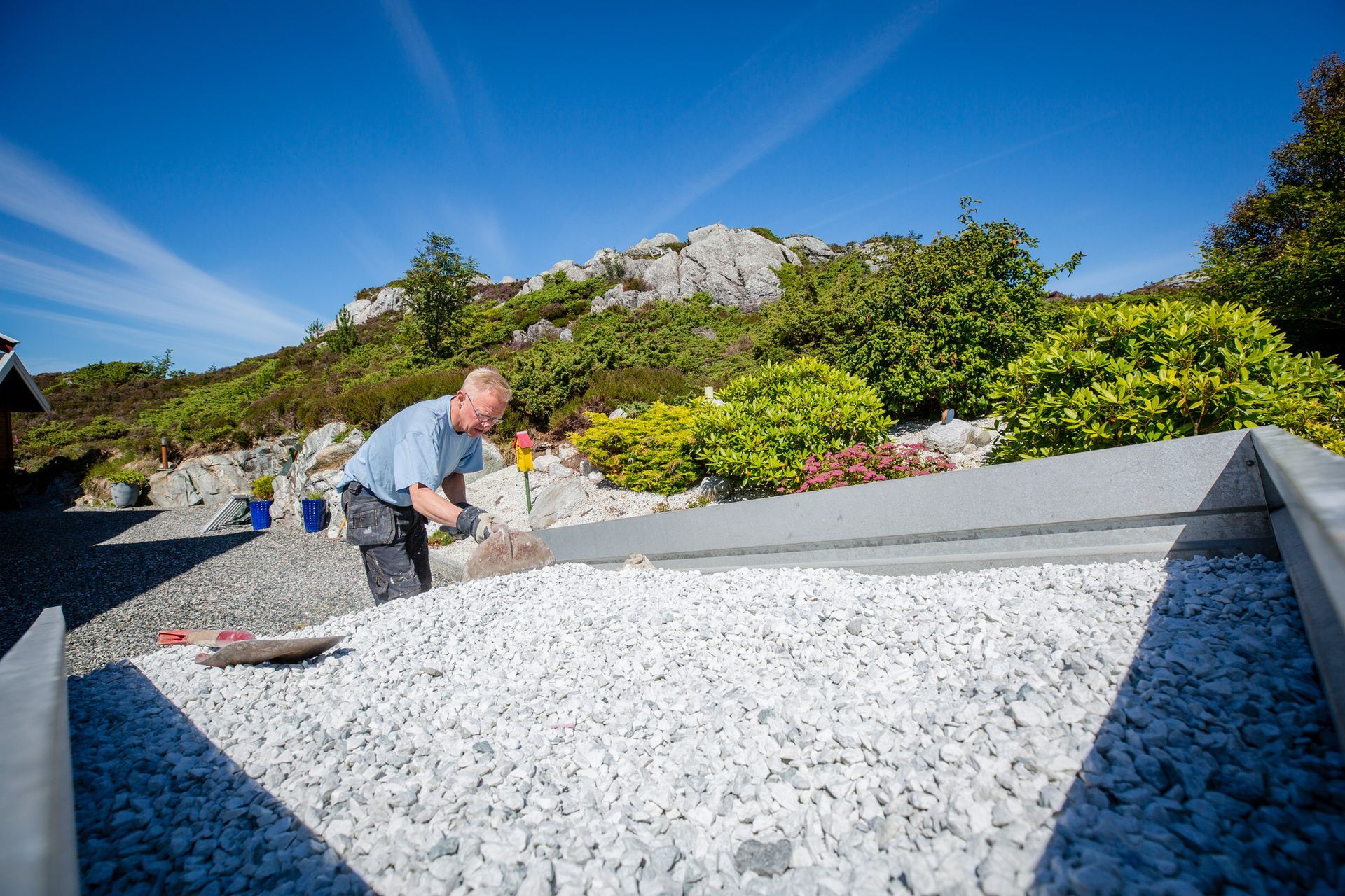 Man spreading gravel in a raised garden bed with a mountain backdrop under a blue sky.