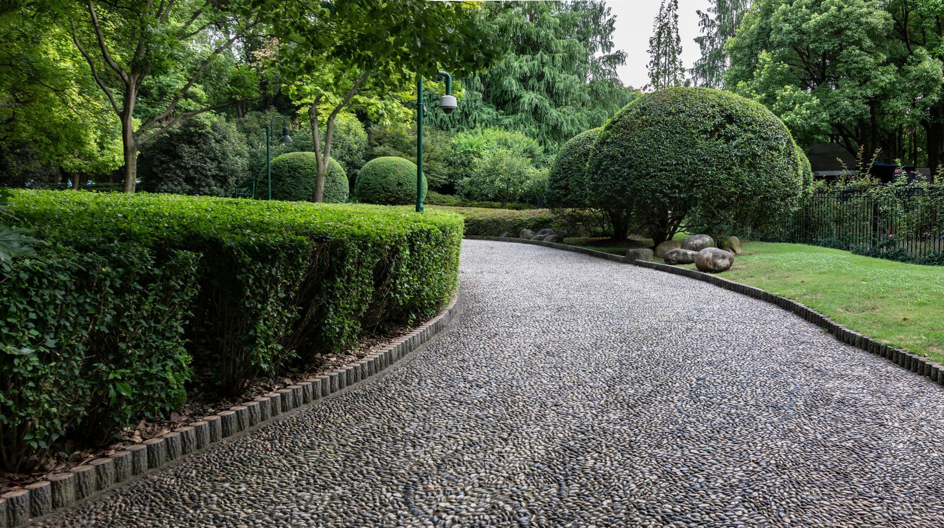 A pebble-covered path curves through a green, landscaped garden with trimmed hedges and trees.