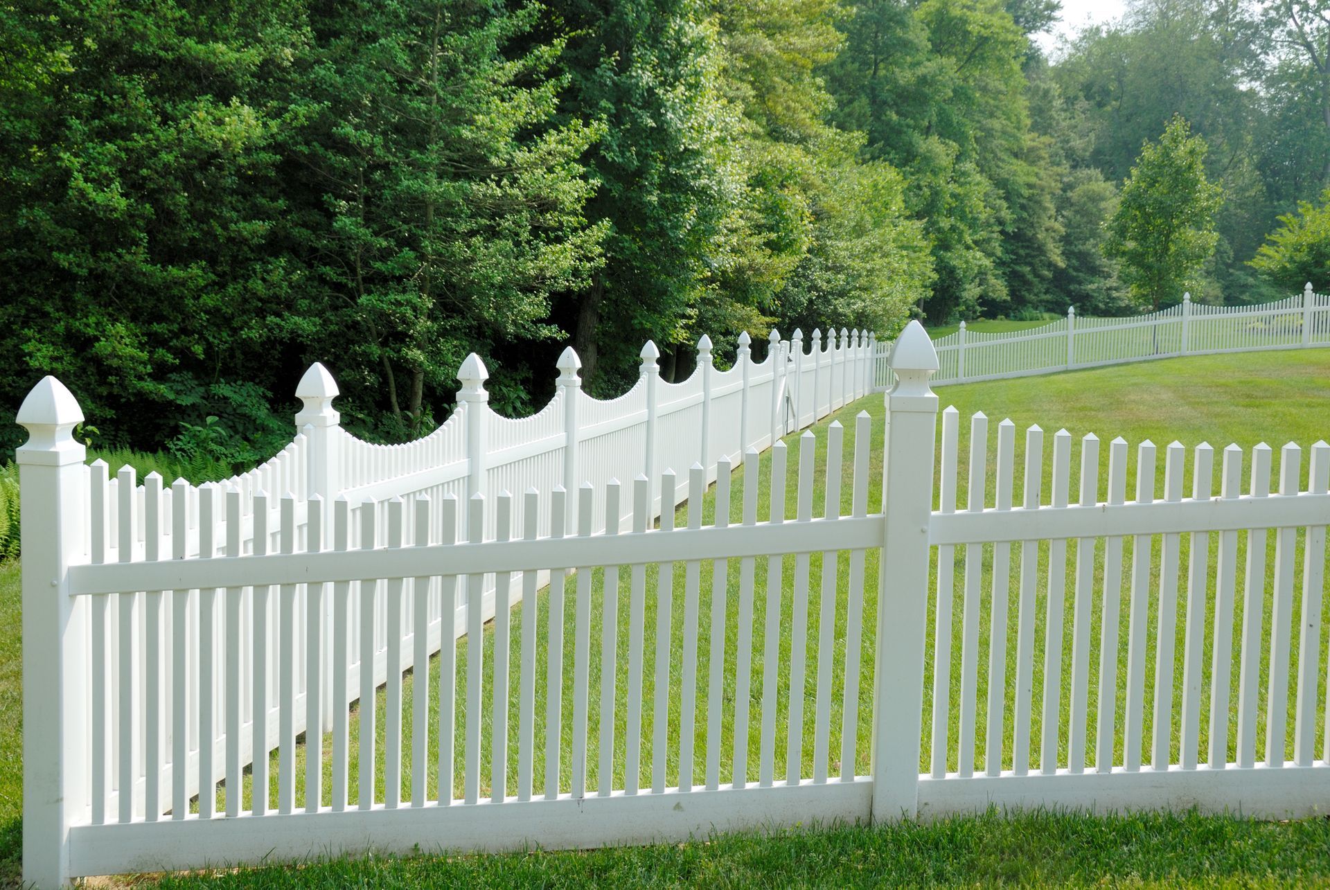 White picket fence bordering a green lawn, with trees in the background.