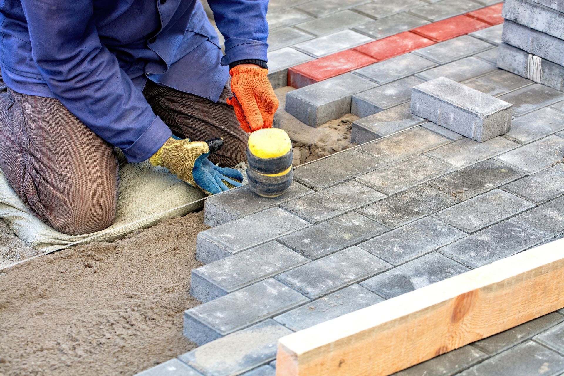 Person laying gray paving stones, using a mallet, on sand, with wood border and some red bricks.