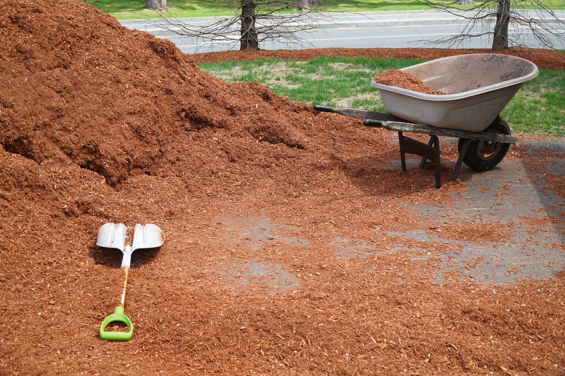 Pile of brown mulch with a wheelbarrow and shovel on a concrete surface, near grass and trees.