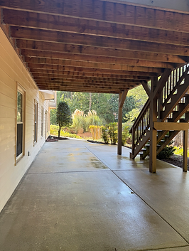 Concrete patio under a wooden deck with stairs. Backyard view with trees and plants.