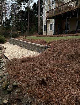 A lakeside yard with a stone retaining wall, beige gravel path, and house with a deck. Pine needles cover the ground.