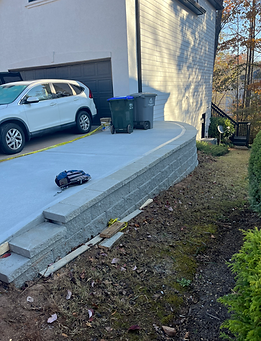 Concrete driveway with a retaining wall, a white SUV, and trash bins.