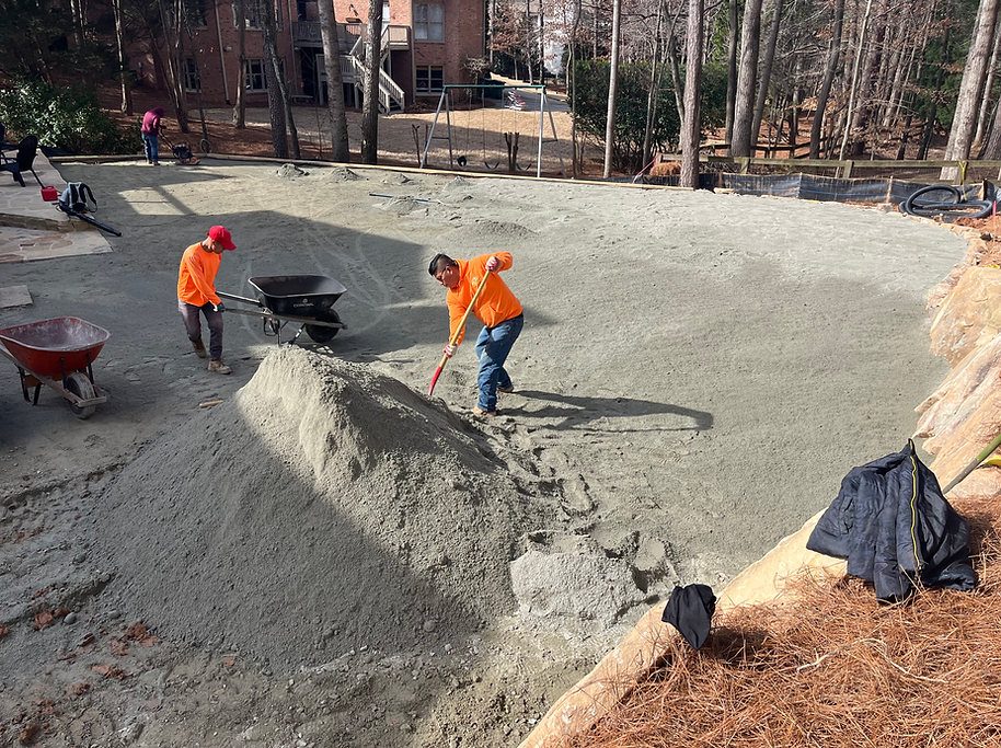 Construction workers spreading concrete in an outdoor area, using shovels and wheelbarrows.