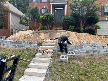 Man building retaining wall and steps in front yard; dirt pile in background.