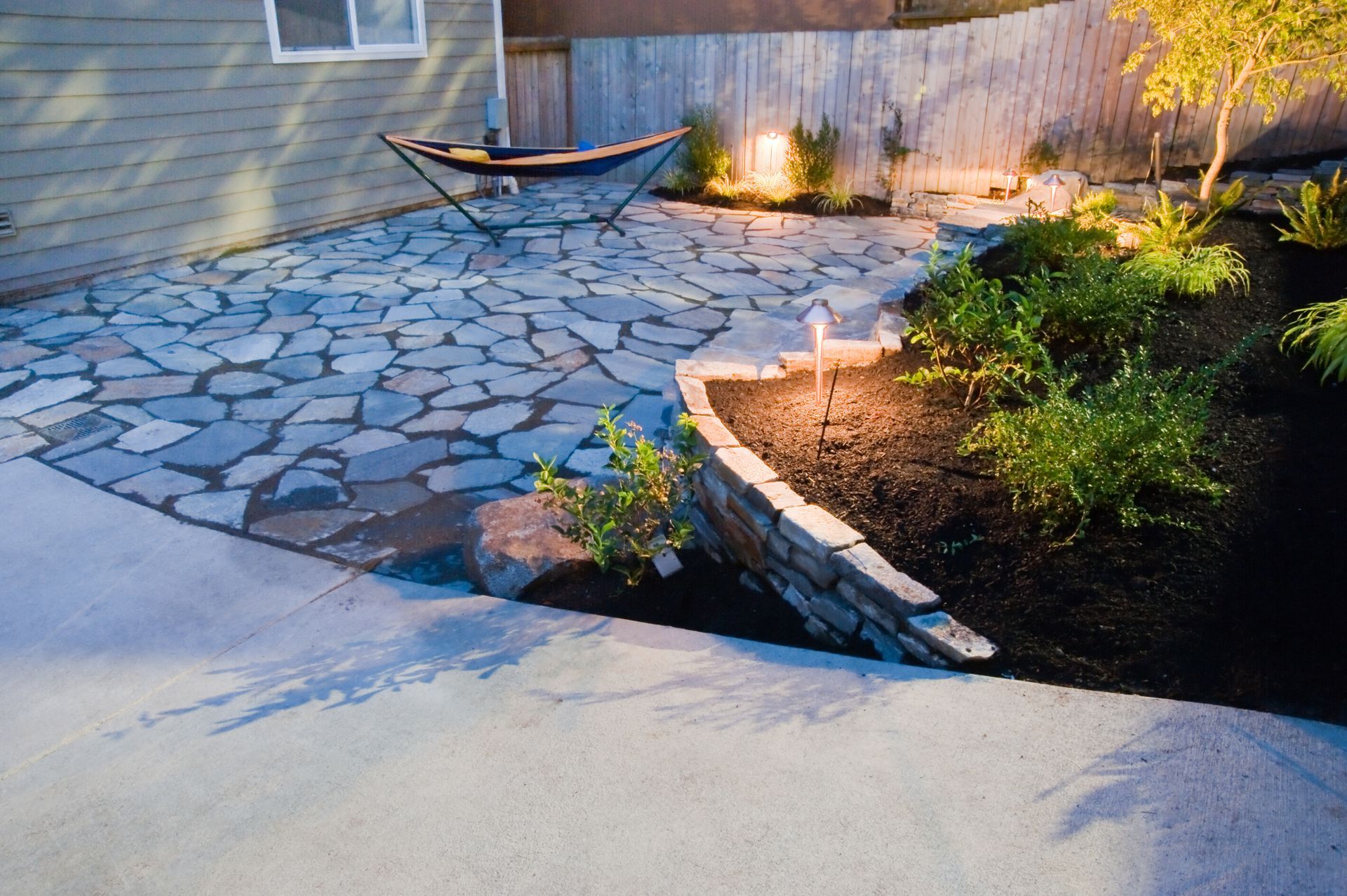 Patio with flagstone pavers, retaining wall, and hammock, illuminated by outdoor lights.