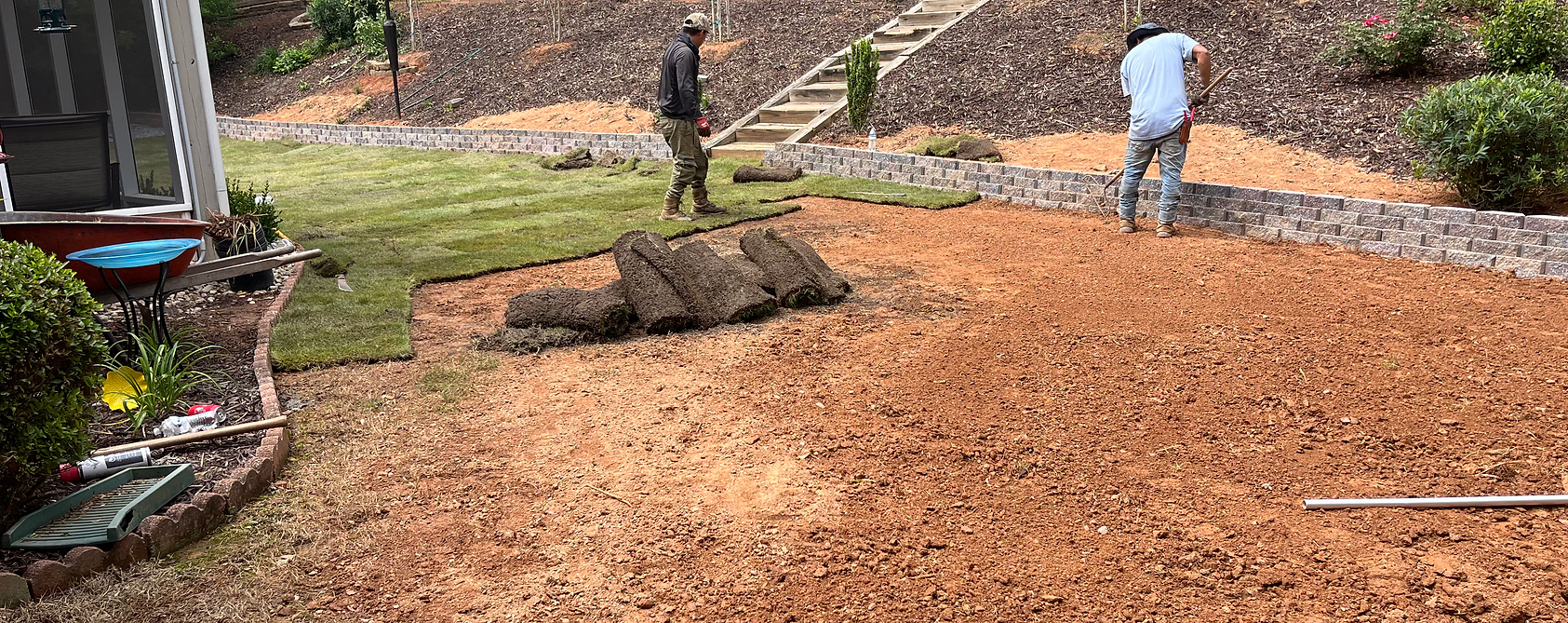 Workers laying sod in a yard. One prepares the soil, another places the sod. Reddish dirt surrounds the green grass.