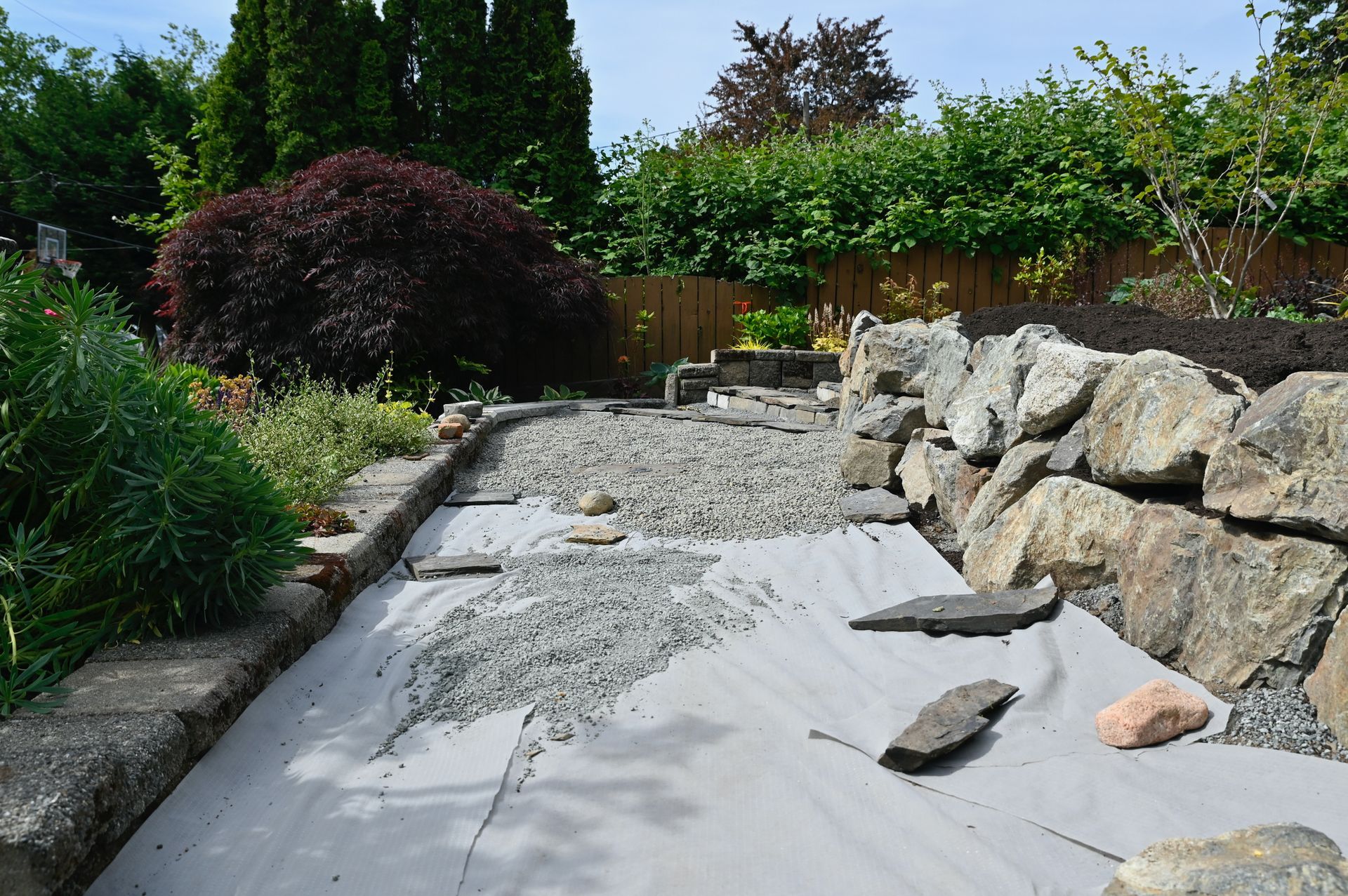 A gravel-filled garden bed with a grey liner, bordered by stone walls and greenery.