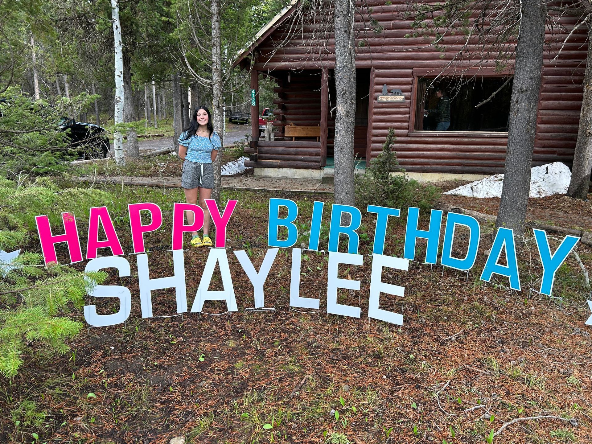 A woman is standing in front of a sign that says `` happy birthday shaylee ''. A woman is standing in front of a sign that says `` happy birthday shaylee ''.