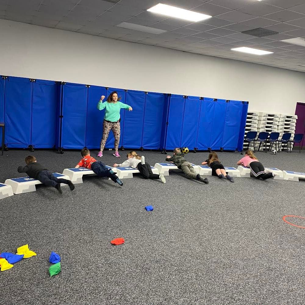 A group of children are laying on the floor in a gym while a woman teaches them
