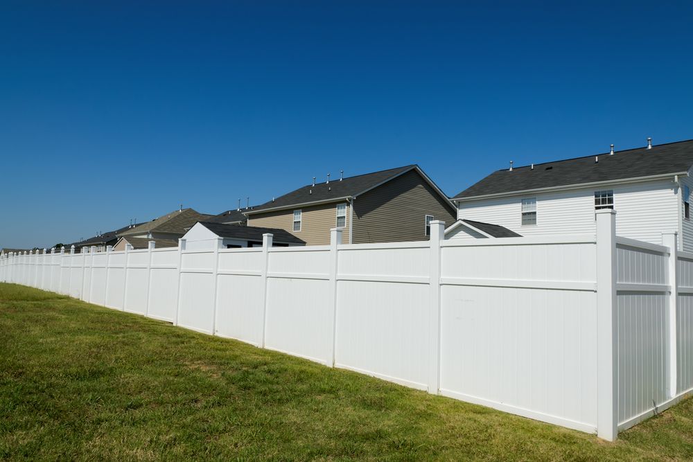 A white fence surrounds a lush green field in front of a row of houses.