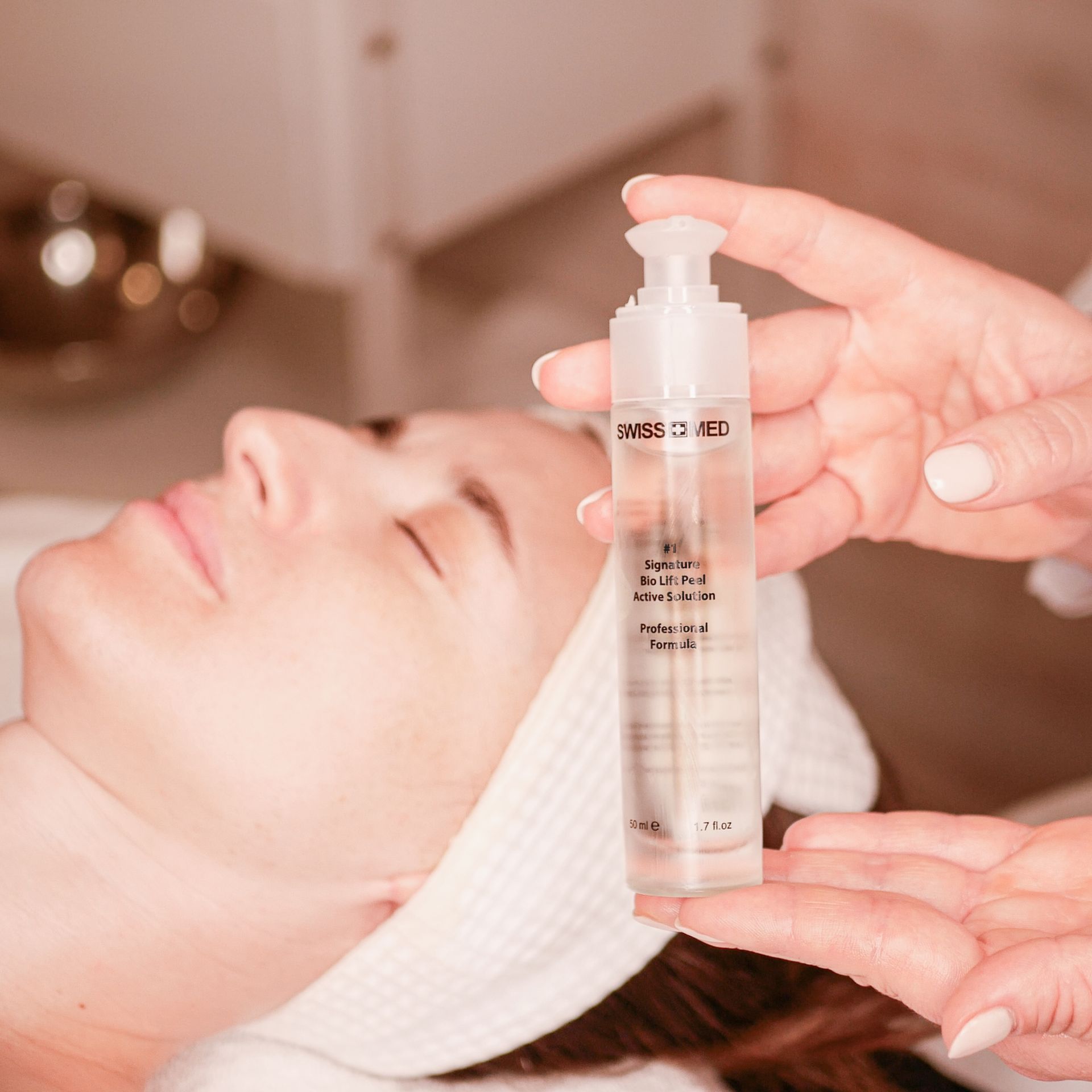 A woman receives facial treatment. Hands hold a skincare product near her face in a spa-like setting.