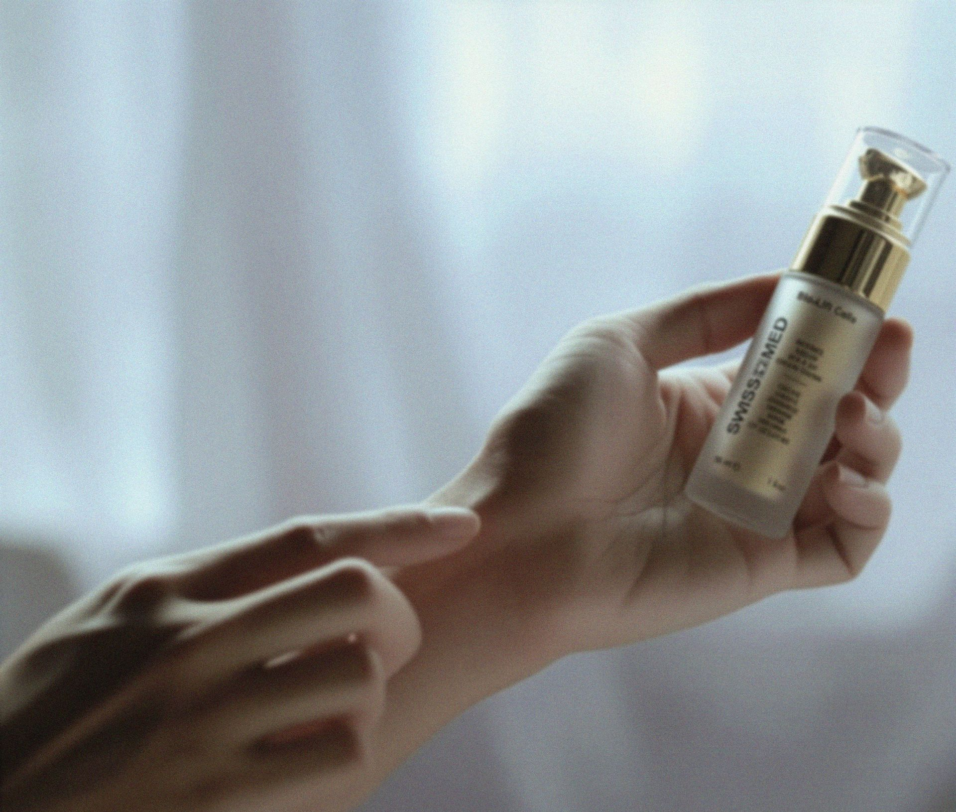 Person applying skincare serum from a gold-capped bottle to their wrist against a neutral background.