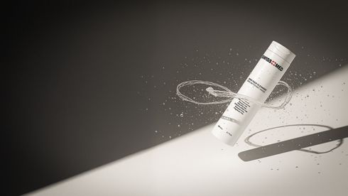 White cylindrical product in mid-air, surrounded by scattering particles, against a two-tone grayscale backdrop.