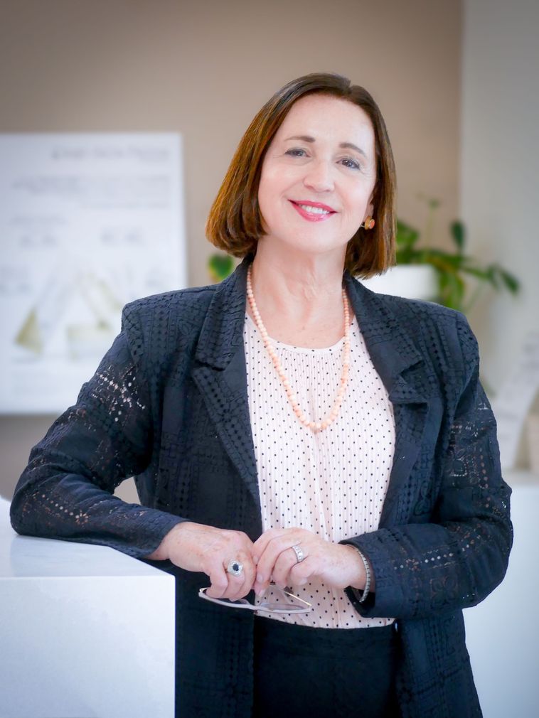 Woman in blazer smiling, leaning on white surface, wearing pearl necklace.
