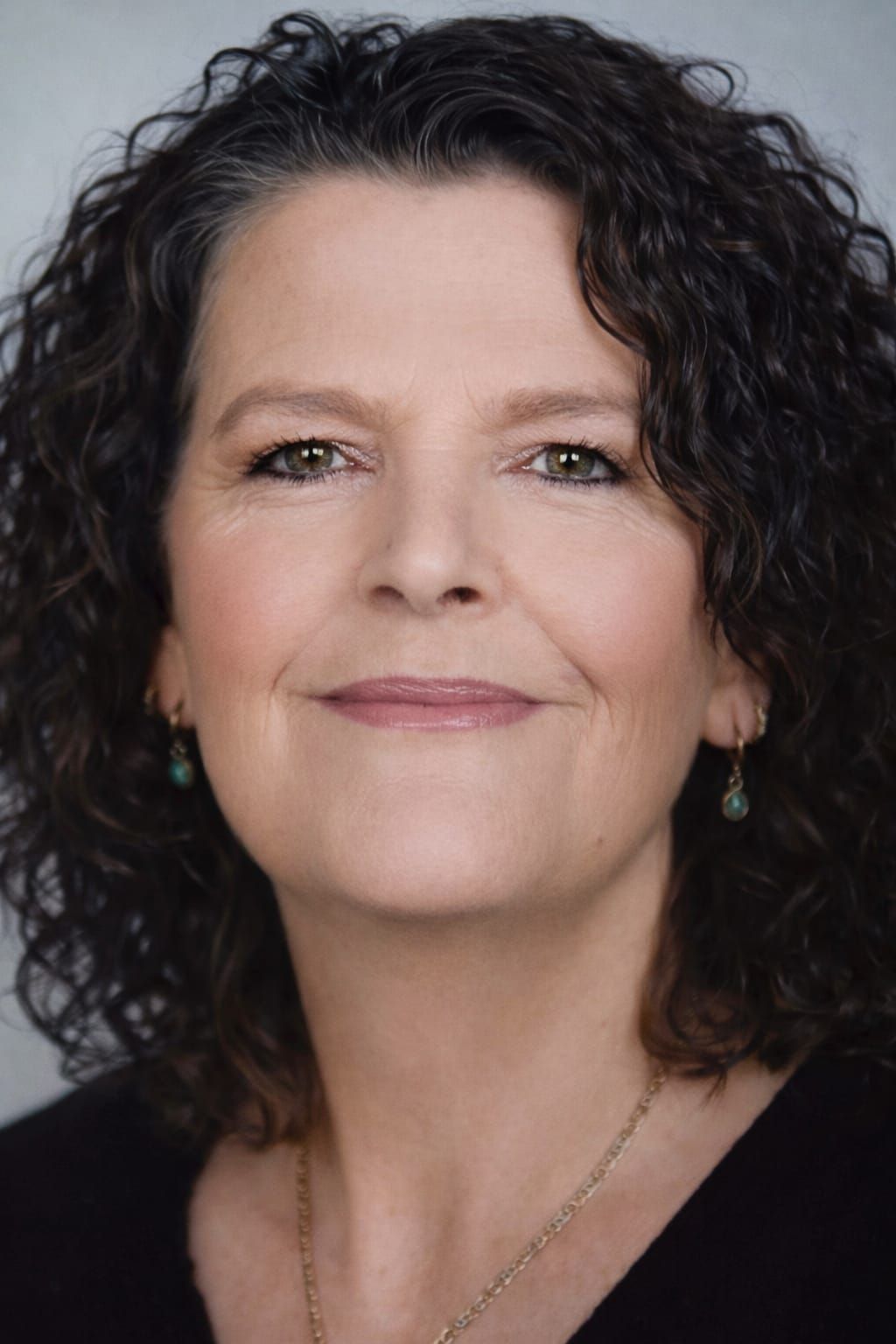 Woman with curly dark hair, smiling, wearing a dark top, with a neutral background.