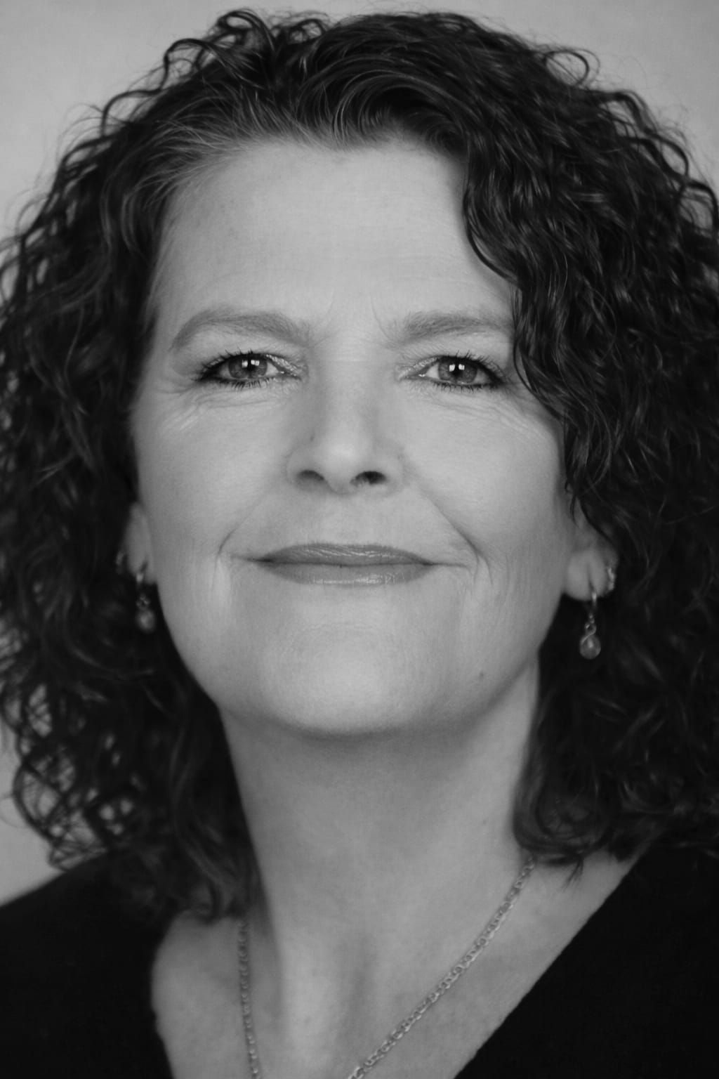 Vrouw met krullend haar lacht en kijkt in de camera. Zwart-witfoto.