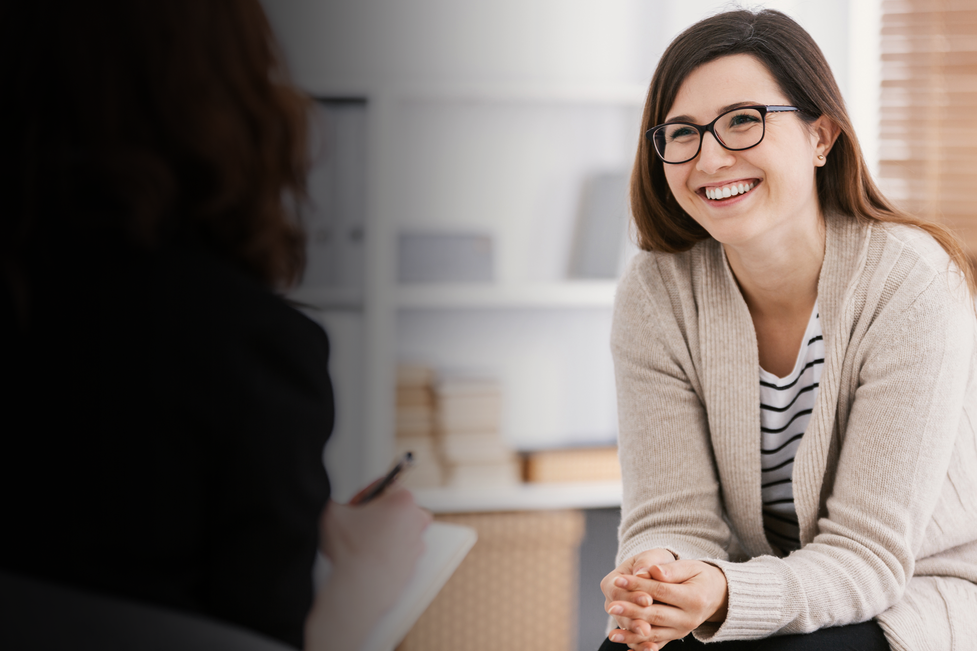 A woman wearing glasses is smiling while talking to another woman.