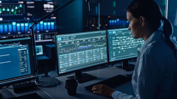 A woman is sitting at a desk in front of three computer monitors.