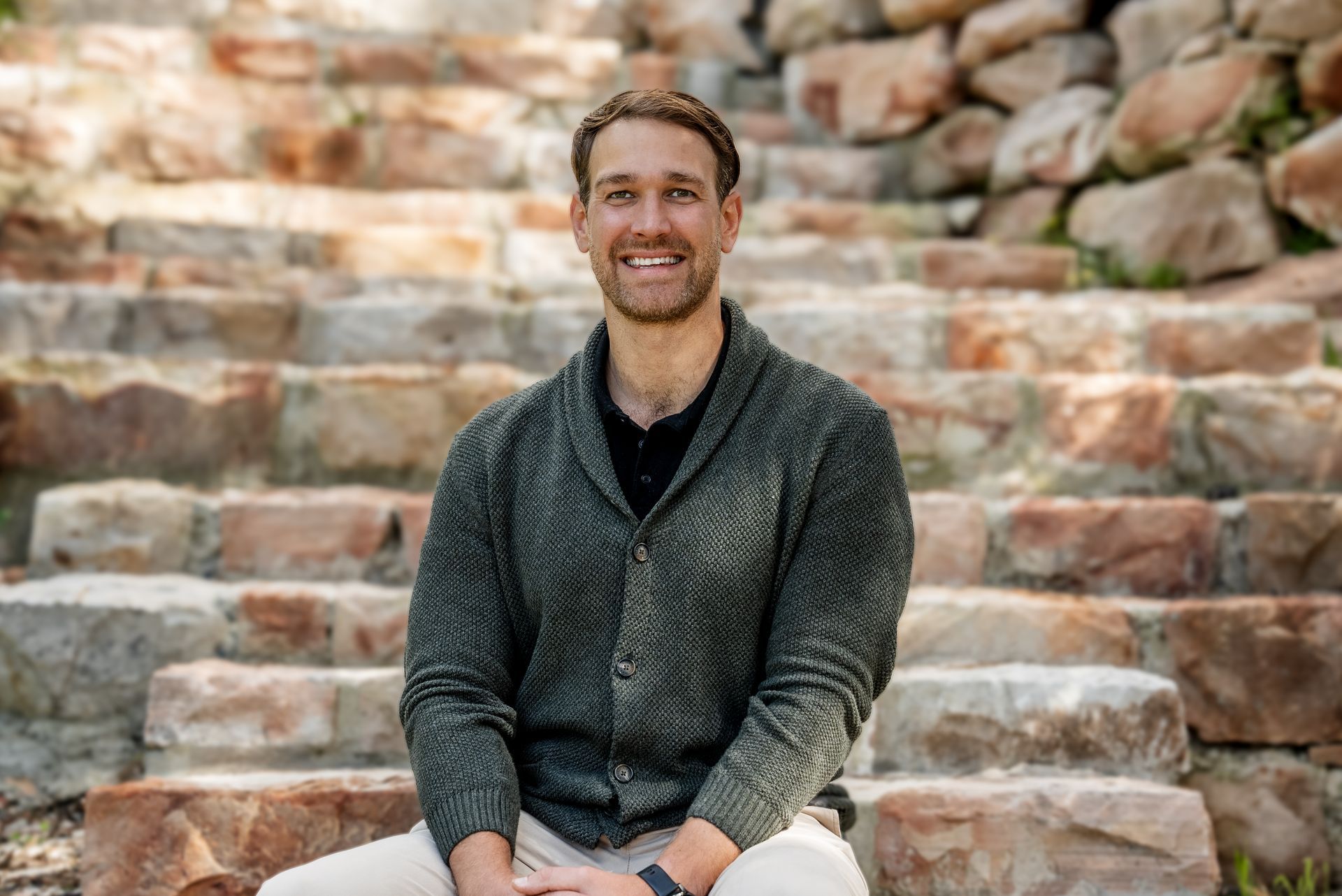 Man in green sweater smiling, sitting on stone steps outdoors.